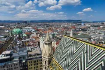 Luftaufnahme einer Stadtlandschaft mit einer markanten Kathedrale mit einem bunten, gemusterten Dach im Vordergrund. Umliegende Geb&auml;ude und entfernte H&uuml;gel sind unter einem teilweise bew&ouml;lkten Himmel sichtbar.