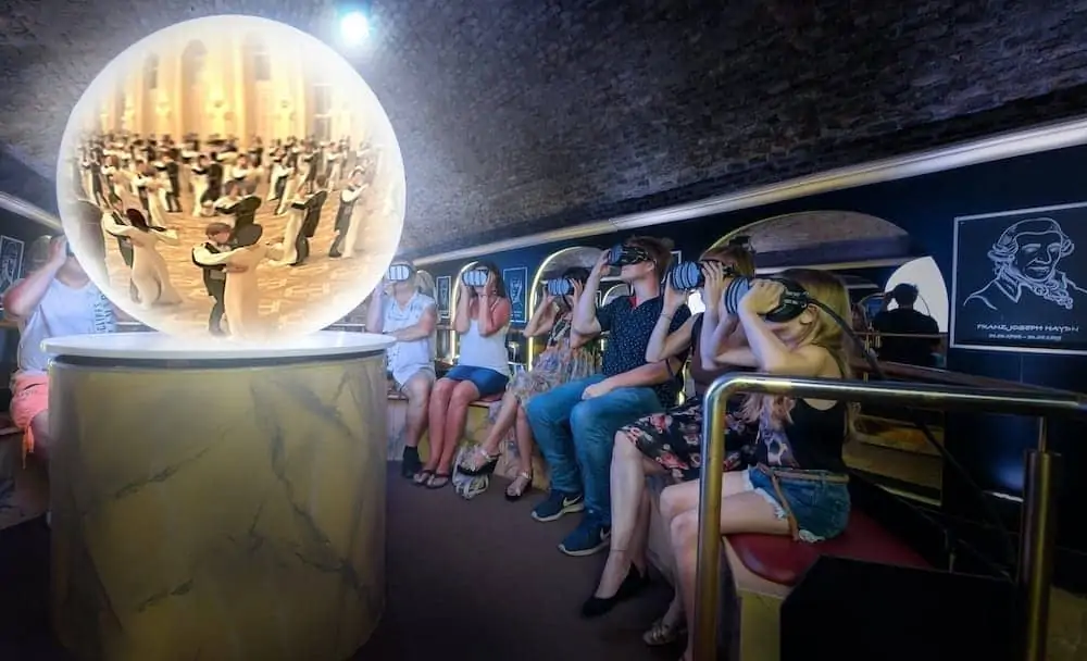 Ludzie siedzą w pokoju i używają zestaw&oacute;w słuchawkowych wirtualnej rzeczywistości. Duża, świecąca kula przed nimi pokazuje scenę tańczącej pary, otoczonej przez widz&oacute;w w strojach wieczorowych. Tło jest nowoczesne i delikatnie oświetlone.  