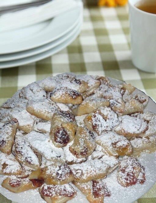 市松模様のテーブルクロスの上に、アイシングシュガーとフルーツスプレッドを添えたウィーン風ポルスタージップが皿いっぱいに盛られている。背景には、積み重ねられた白い皿、カトラリー、ひまわり、紅茶のカップが置かれ、魅力的なシーンが完成している。