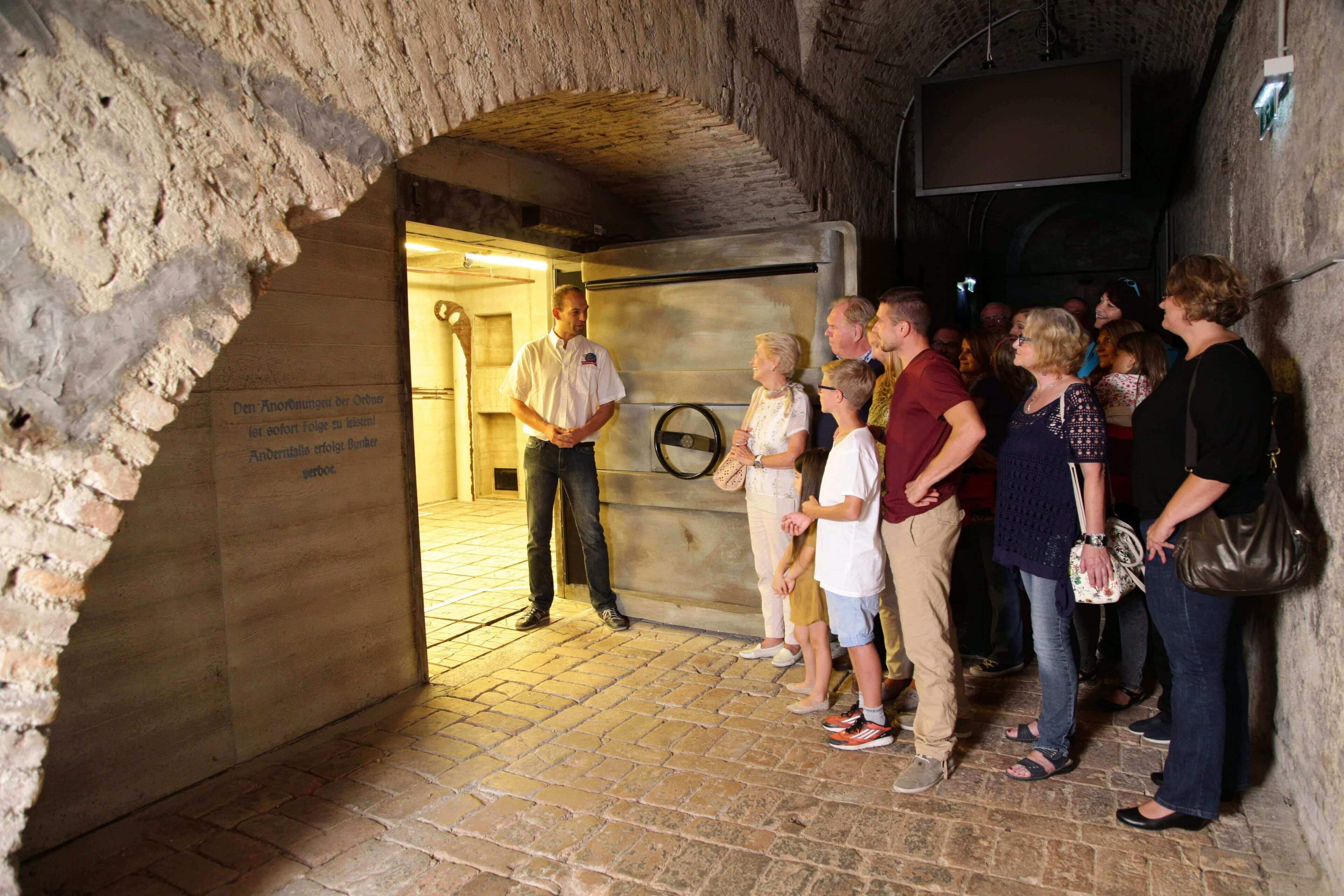 Un guía turístico está de pie junto a una gran puerta metálica en una sala subterránea con paredes de ladrillo y explica algo a un grupo de adultos y niños que escuchan atentamente.