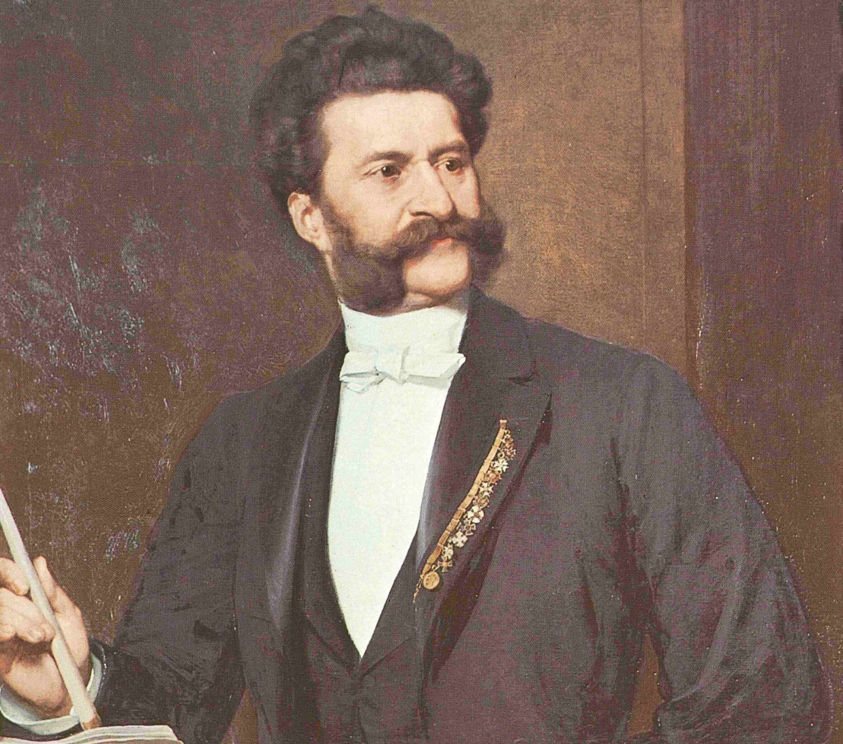 ヨハン・シュトラウス-音楽革命黒髪に大きなもみあげの男性が、フォーマルな黒いスーツに白いシャツと蝶ネクタイを身に着けている。タクトを手に指揮台に立ち、聴衆に向かって指揮かスピーチをしているように見える。