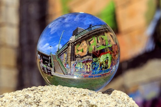 Una sfera di vetro poggia su una roccia e riflette un muro ricoperto di graffiti colorati, una scala e il cielo blu di una vicina area urbana. I graffiti e l'edificio appaiono capovolti nella sfera di cristallo.