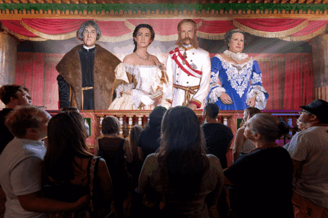 一群人看着四尊身着精致历史服饰、栩栩如生的雕像站在红色幕布和戏剧性灯光的剧院舞台上。