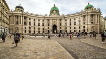 Emberek s&eacute;t&aacute;lnak a macskak&ouml;ves t&eacute;ren a Hofburg-palota előtt B&eacute;csben, Ausztri&aacute;ban. A t&ouml;rt&eacute;nelmi barokk &eacute;p&uuml;let nagy &iacute;ves homlokzata &eacute;s z&ouml;ld kupol&aacute;s teteje felhős &eacute;gbolt alatt.