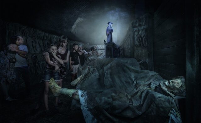 Un gruppo di persone sembra terrorizzato mentre cammina accanto a inquietanti cadaveri in decomposizione in un buio tunnel sotterraneo. Alla fine del tunnel, un'oscura figura con un cappello si trova sotto un fascio di luce, contribuendo ad aumentare l'atmosfera inquietante.