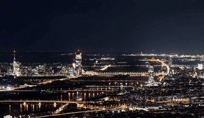 Paesaggio urbano notturno con edifici illuminati, ponti e lampioni riflessi in un fiume. Lo skyline è costituito da un mix di edifici alti e bassi sotto un cielo scuro.