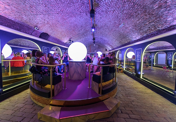 Eine Gruppe von Menschen sitzt in einem kreisförmigen Raum unter einem Ziegelgewölbe um eine große leuchtende Kugel herum. Der Raum ist mit violetten und blauen Lichtern beleuchtet, die Wände sind verspiegelt und an den Seiten befinden sich gewölbte Nischen.