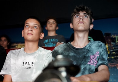 Eine Gruppe von Jungen sitzt eng beieinander in einem dunklen Raum und blickt mit konzentriertem Blick nach oben, der von oben beleuchtet wird. Die beiden Jungen im Vordergrund tragen Krawattenhemden und wirken verlobt.