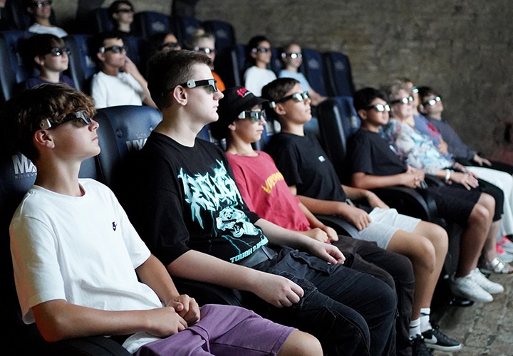 Eine Gruppe junger Leute mit 3D-Brillen sitzt in Kinosesseln und verfolgt aufmerksam einen Film oder eine Präsentation auf einer großen Leinwand. Der Raum ist schwach beleuchtet, was ihre konzentrierte Mimik unterstreicht.