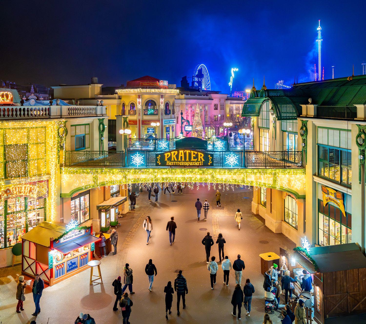 Eine festliche Nachtszene im Wiener Prater während des Wintermarkts, mit Menschen, die unter einem geschmückten Torbogen spazieren gehen, beleuchteten Gebäuden, funkelnden Lichtern und einem Riesenrad im Hintergrund.