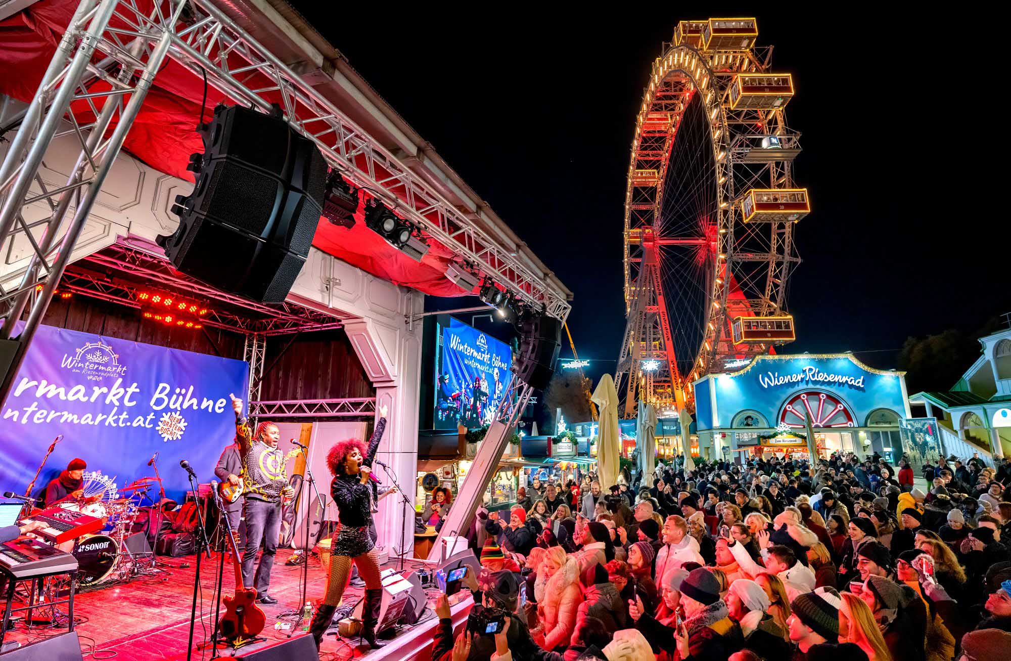Ein lebhaftes Konzert unter freiem Himmel bei Nacht mit einem großen Publikum, einer Band auf der Bühne, hellen Lichtern und einem beleuchteten Riesenrad im Hintergrund in einem Vergnügungspark während des Wintermarkts.