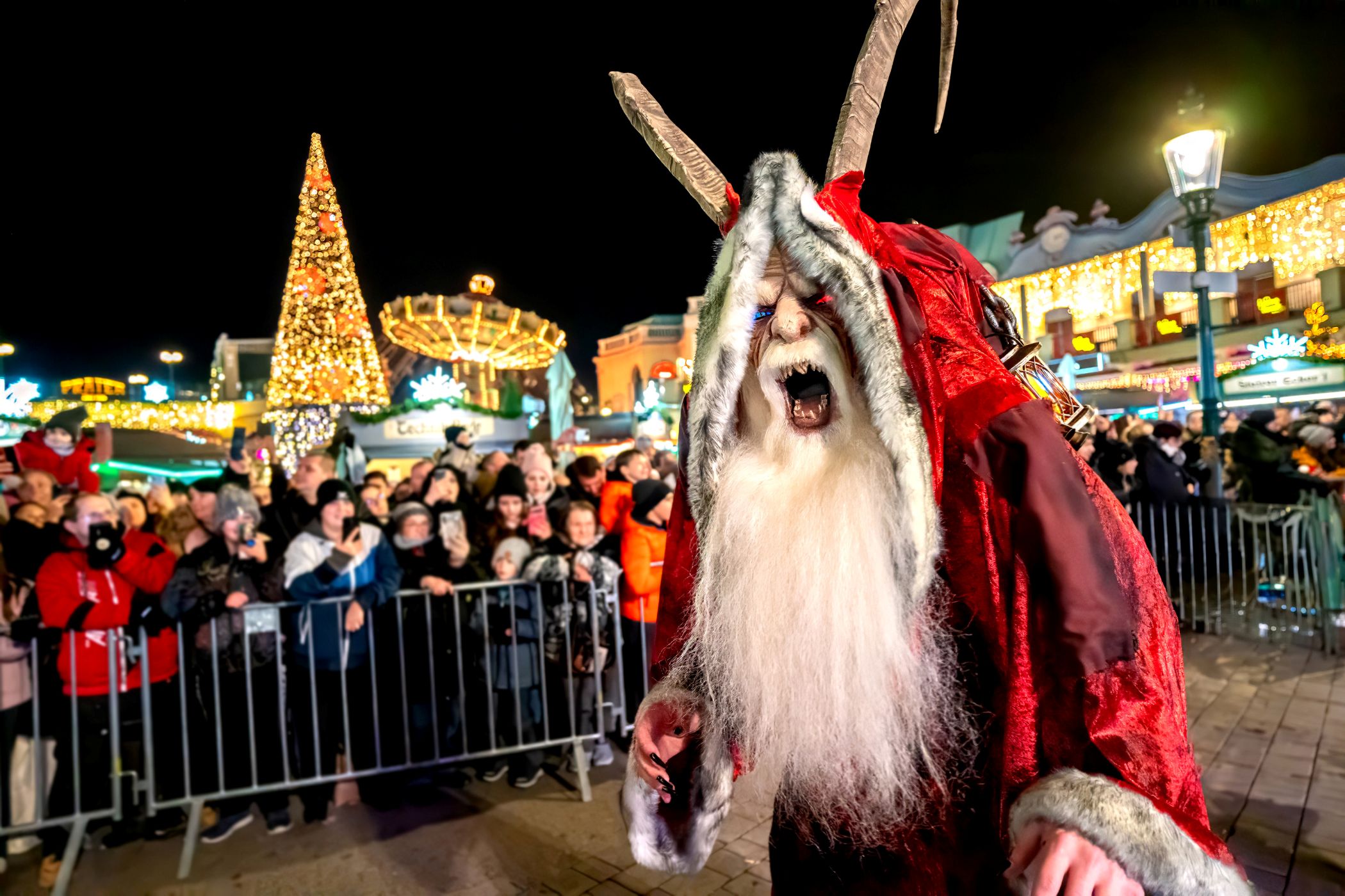 Eine Person in einem roten Krampuskostüm mit großen Hörnern und weißem Bart geht nachts in der Nähe einer festlichen Wintermarktmenge spazieren. Im Hintergrund leuchten Weihnachtslichter und ein geschmückter Baum.