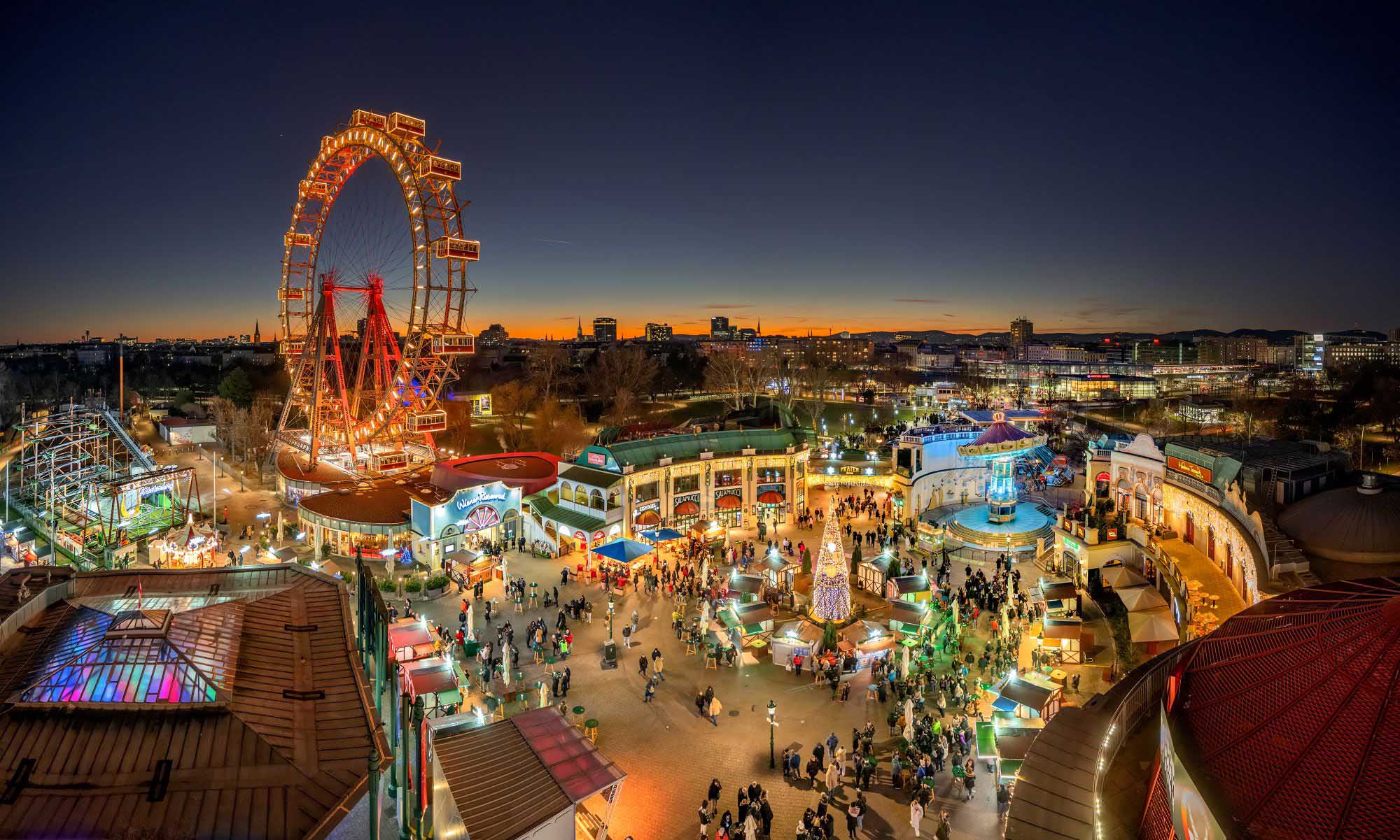 Ein belebter Wintermarkt in der Abenddämmerung mit einem großen beleuchteten Riesenrad, bunten Ständen, Menschenmassen und Lichtern der Stadt, die unter einem tiefblauen und orangefarbenen Abendhimmel leuchten.