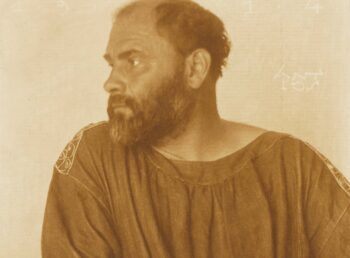 Sepiafarbenes Porträt von Gustav Klimt in einem lockeren, bestickten Hemd, der mit ernster Miene nach links schaut, vor einem schlichten Hintergrund. Im Hintergrund erscheinen schwache Zahlen und Buchstaben.