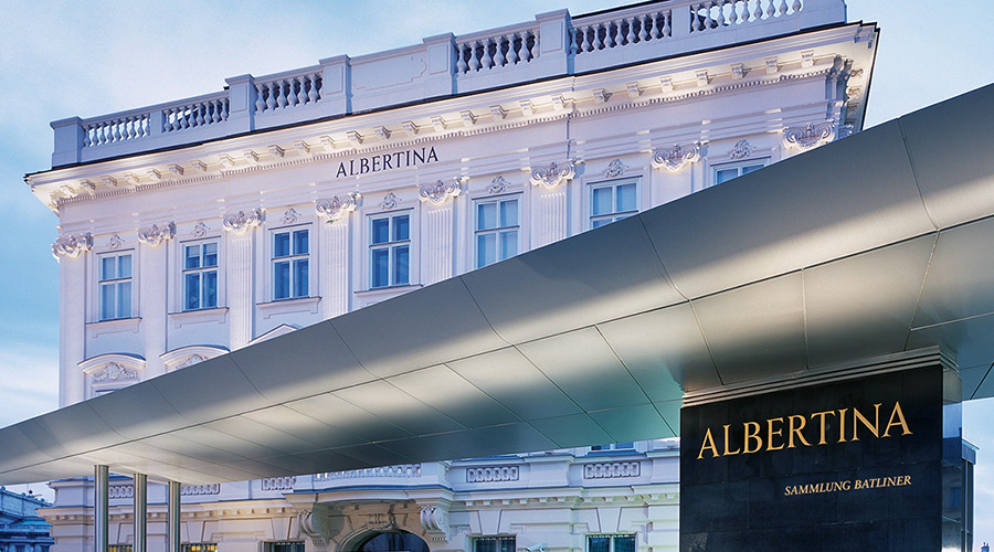 一座宏伟的白色历史建筑,上面刻有 ALBERTINA 字样,并有华丽的细节装饰。前景是一座现代建筑和一个黑色标志,上面也写着阿尔伯蒂纳。天空晴朗,呈淡蓝色。