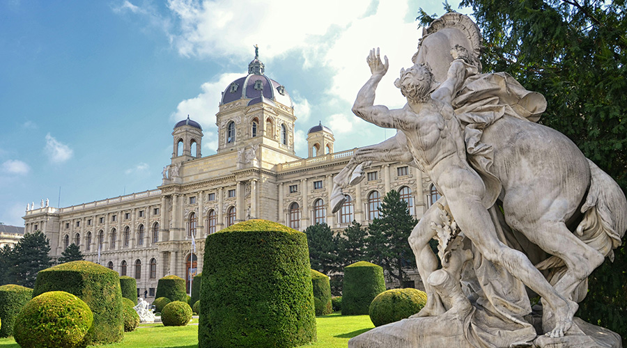 在明亮的蓝天下,一座大型历史建筑矗立在修剪整齐的花园和引人注目的大理石雕像后面,雕像的中央是圆顶和拱形窗户。