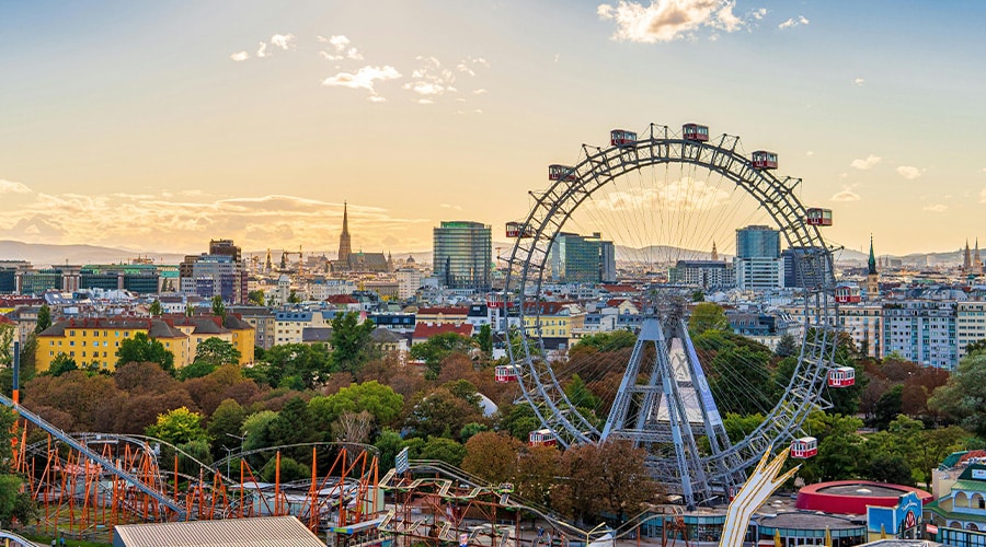 遊園地に立つ大観覧車とジェットコースター。背景には夕暮れ時の一部曇り空の下、街並みと教会の尖塔。