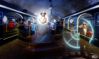 Eine Gruppe von Menschen betrachtet in einem tunnelartigen Raum eine große beleuchtete Beethoven-Büste; digitale Lichteffekte wirbeln herum, und ein Mann mit Sonnenbrille zeigt auf eine Fernbedienung, was eine surreale, moderne Atmosphäre schafft.