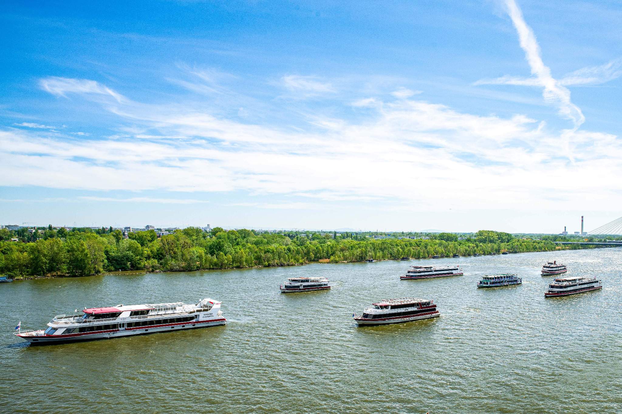 Mehrere weiße und rote Boote fahren auf einem breiten Fluss unter einem blauen Himmel mit Wolkenfetzen. Grüne Bäume säumen das Flussufer, und in der Ferne ist rechts eine Brücke zu sehen.