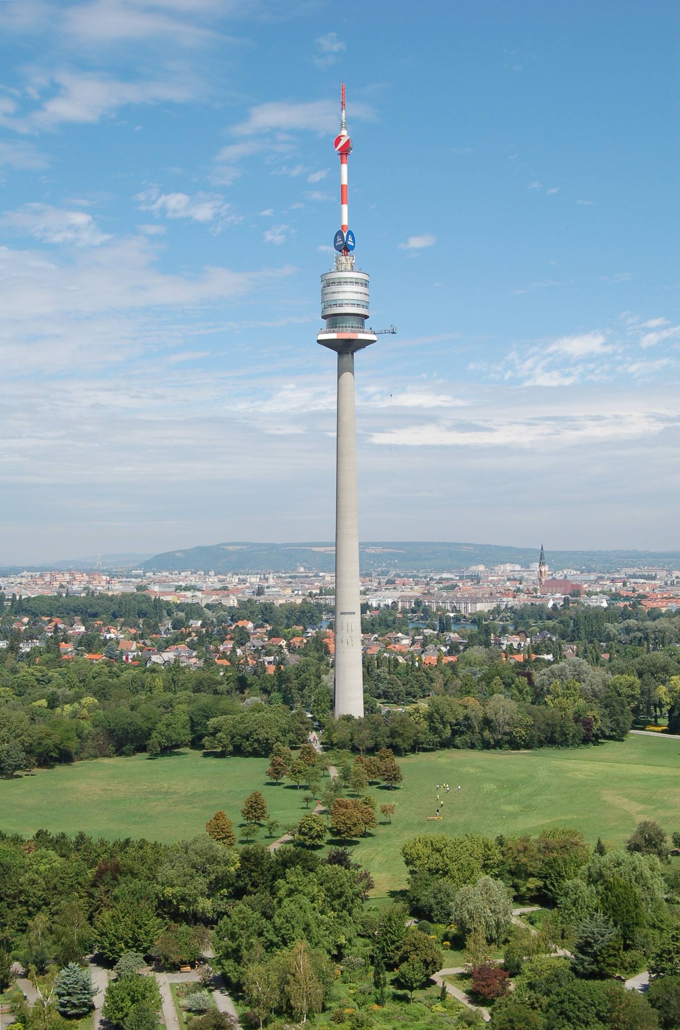 Ein hoher Fernmeldeturm mit einer rot-weißen Antenne erhebt sich über einem grünen Park, in dem eine Stadtlandschaft und entfernte Hügel unter einem teilweise bewölkten Himmel zu sehen sind.