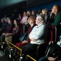 Eine bunt gemischte Gruppe Erwachsener sitzt in Reihenbestuhlung im Theater und verfolgt aufmerksam eine Show oder Präsentation mit lächelnden und konzentrierten Gesichtern bei gedämpftem Licht.