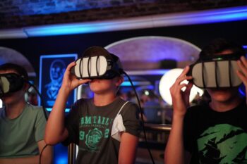 Drei Kinder sitzen mit Virtual-Reality-Headsets nebeneinander in einem schwach beleuchteten Raum mit blauen und violetten Lichtern und scheinen mit der VR-Erfahrung besch&auml;ftigt zu sein.