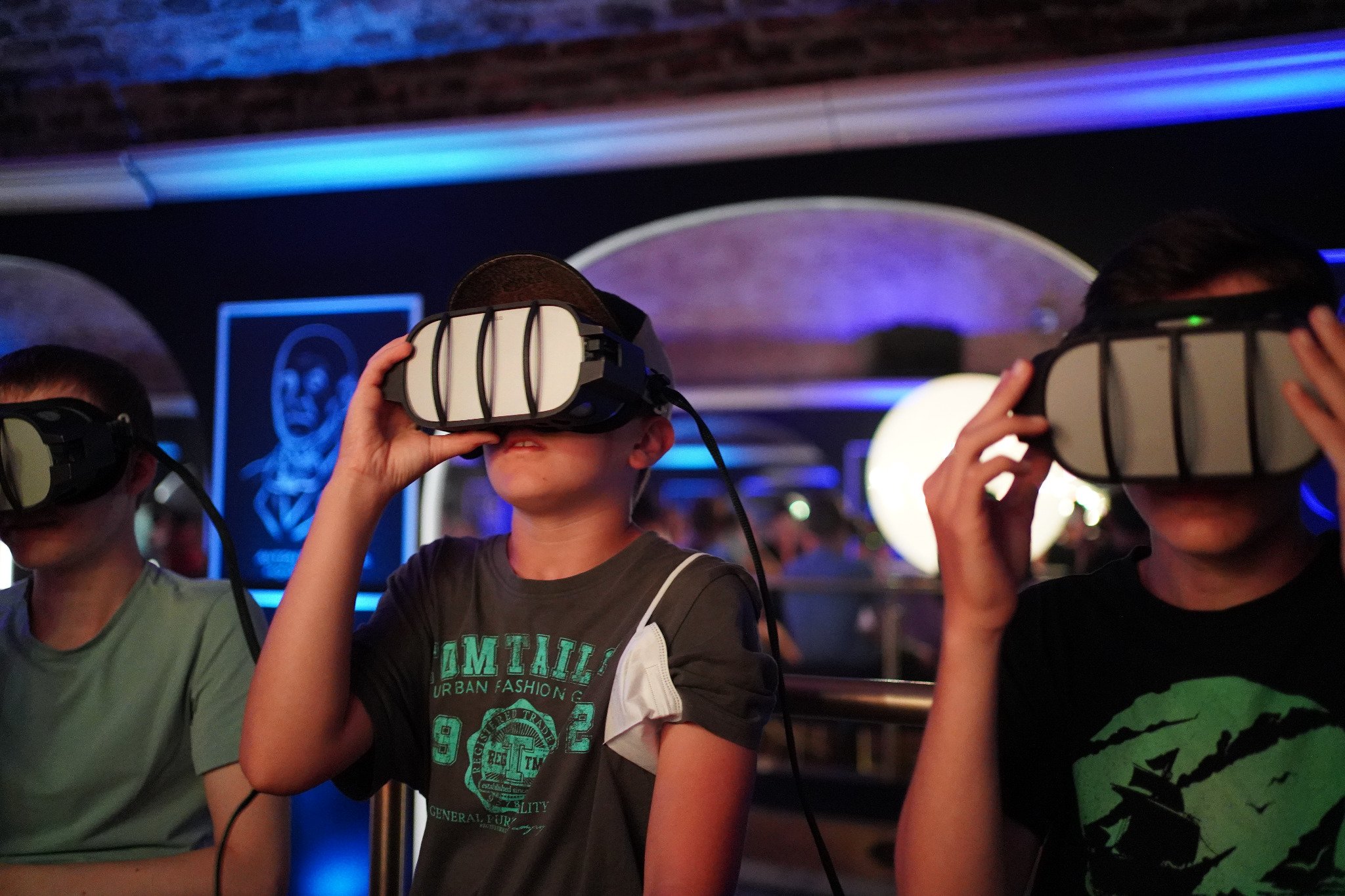Drei Kinder sitzen mit Virtual-Reality-Headsets nebeneinander in einem schwach beleuchteten Raum mit blauen und violetten Lichtern und scheinen mit der VR-Erfahrung beschäftigt zu sein.