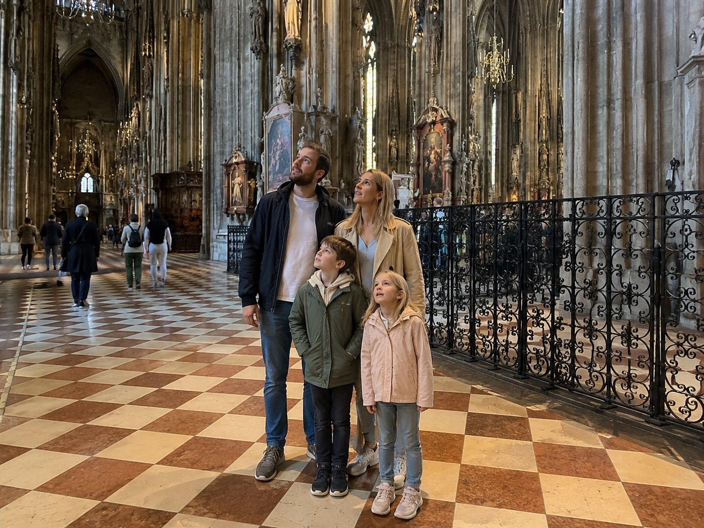 Eine vierköpfige Familie, zwei Erwachsene und zwei Kinder, steht zusammen in einer großen Kathedrale mit hohen Gewölben, verzierten Säulen und schachbrettartigen Böden und betrachtet die Architektur. Im Hintergrund sind weitere Besucher zu sehen.