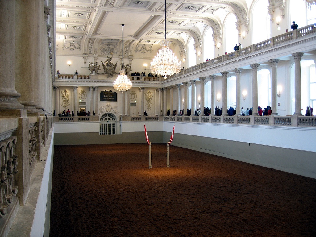 Eine elegante Reithalle mit Kronleuchtern, weißen, verzierten Wänden und hohen Decken. Die Zuschauer schauen von Balkongalerien über einer großen Arena mit brauner Erde und zwei roten Stangen in der Mitte zu.