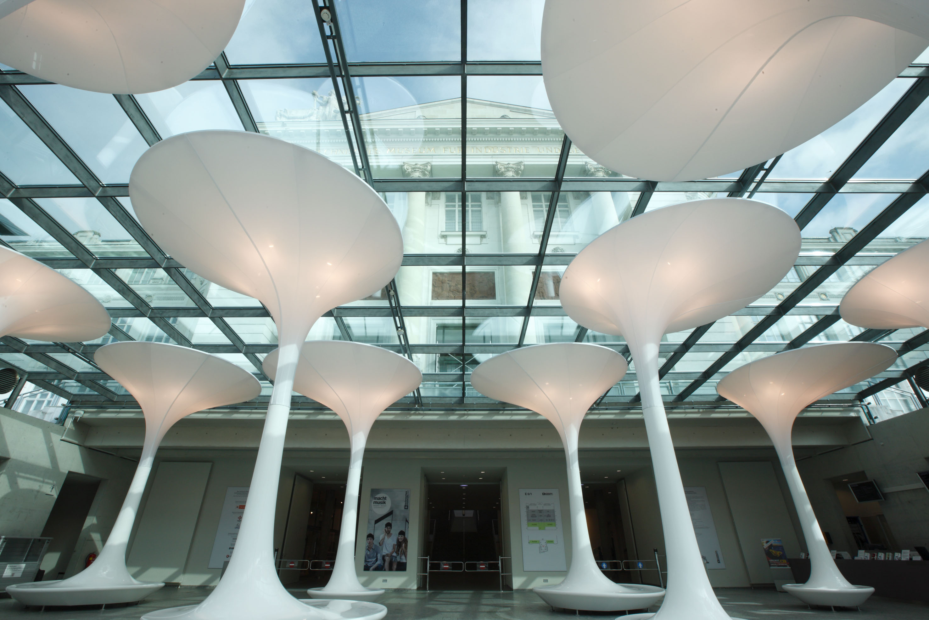 Große, weiße, futuristische pilzförmige Strukturen stehen unter einer Glasdecke in einem modernen Gebäude mit einer klassischen Fassade, die durch das Dach sichtbar ist. Der geräumige, offene Innenraum wird von natürlichem Licht durchflutet.