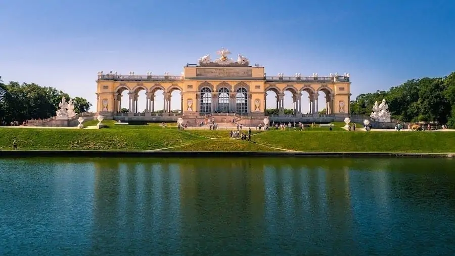Das Foto zeigt die Gloriette, ein gro&szlig;artiges, bogenf&ouml;rmiges Bauwerk mit Skulpturen auf einem grasbewachsenen H&uuml;gel, das sich in einem Teich spiegelt, unter einem klaren blauen Himmel in Wien, &Ouml;sterreich.