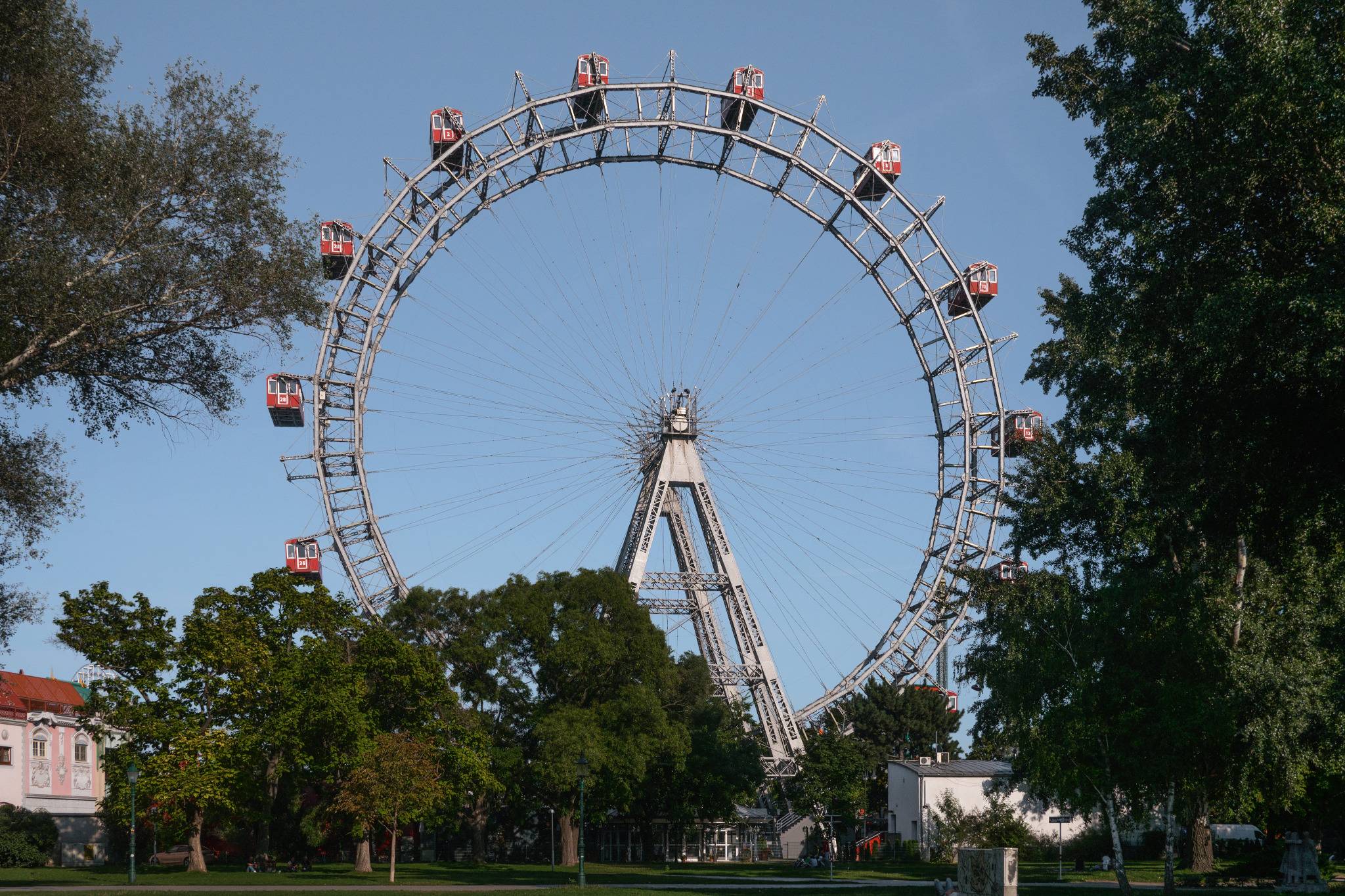 Ein großes Riesenrad mit roten Kabinen steht in einem Park, umgeben von grünen Bäumen und unter einem klaren blauen Himmel.