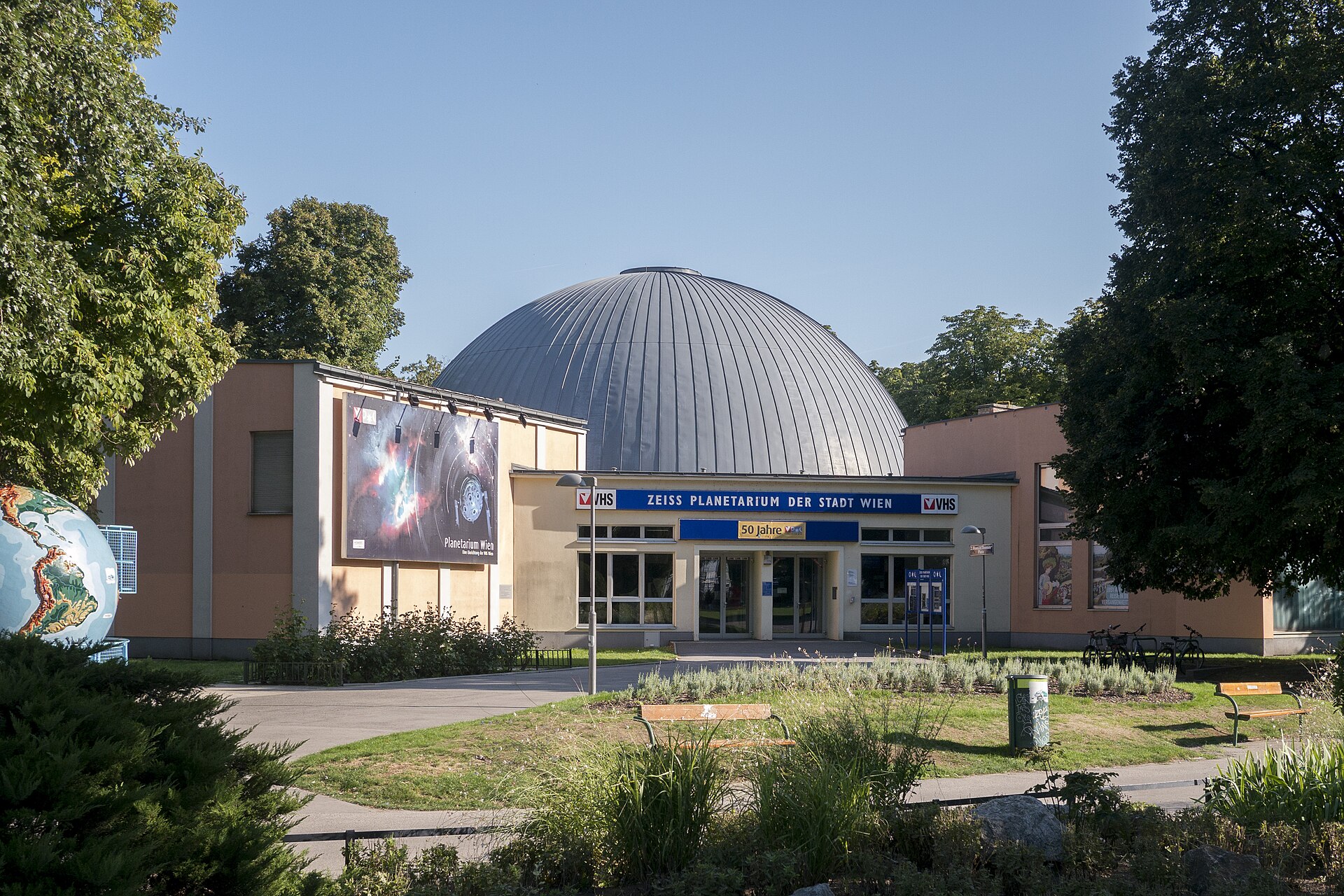 Un gran edificio abovedado, el Planetario Zeiss de Viena, se alza en medio de los árboles, con carteles sobre la entrada y un mural sobre el tema del espacio en la pared izquierda, en un día soleado de cielo despejado.