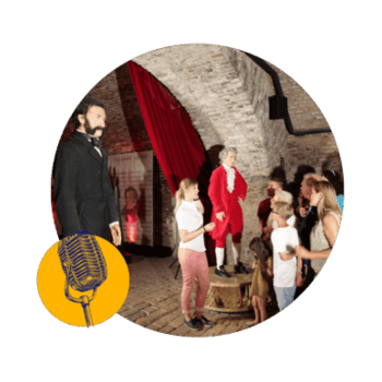 Des personnes, dont des enfants, se rassemblent autour de figures de cire vêtues de costumes d'époque dans une pièce aux murs de pierre, au plafond voûté et aux rideaux rouges. Dans un coin, on aperçoit l'illustration d'un microphone à l'ancienne.