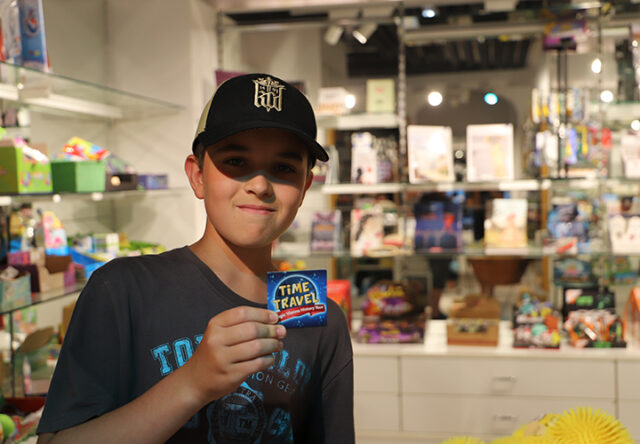 Ein Junge mit schwarzer Mütze und grauem T-Shirt lächelt, während er in einem hell erleuchteten Laden voller Spielzeug und Spiele eine bunte Verpackung mit der Aufschrift „TIME TRAVEL“ in der Hand hält.
