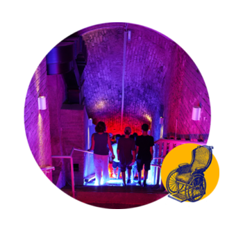 Quatre personnes descendent un escalier dans une pièce aux allures de tunnel, éclairée par des lumières colorées, violettes et bleues ; dans le coin inférieur droit, on aperçoit une petite illustration représentant un vieux fauteuil roulant.
