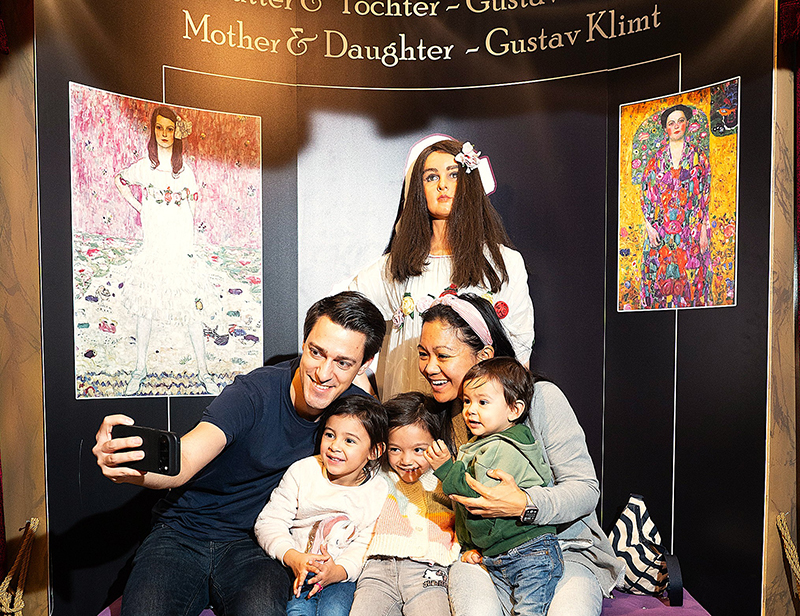 A family of four takes a selfie together in front of an exhibition featuring Gustav Klimt’s “Mother and Daughter,” which includes two large portraits set against floral backgrounds. The family is smiling and sitting close together on a bench.