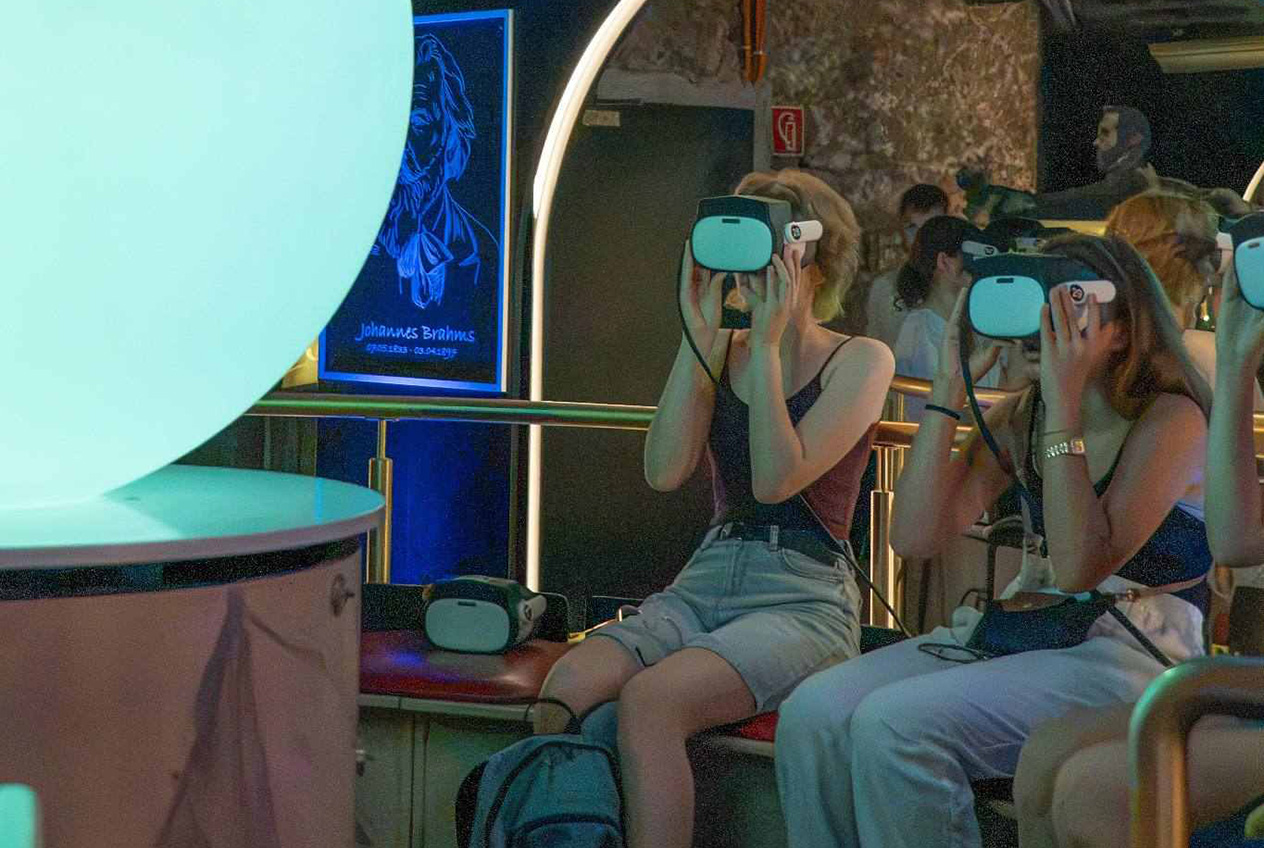 Eine Gruppe von Menschen sitzt in einem Raum, trägt Virtual-Reality-Brillen und blickt auf eine große, leuchtende Kugel. Einige haben Rucksäcke auf dem Schoß. Im Hintergrund ist ein Neonkunstwerk zu sehen.