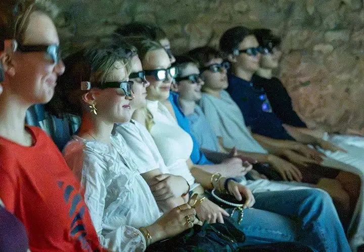 Eine Gruppe von Menschen mit 3D-Brillen sitzt dicht beieinander in einem dunklen Kinosaal und blickt mit konzentrierten Mienen auf die Leinwand.