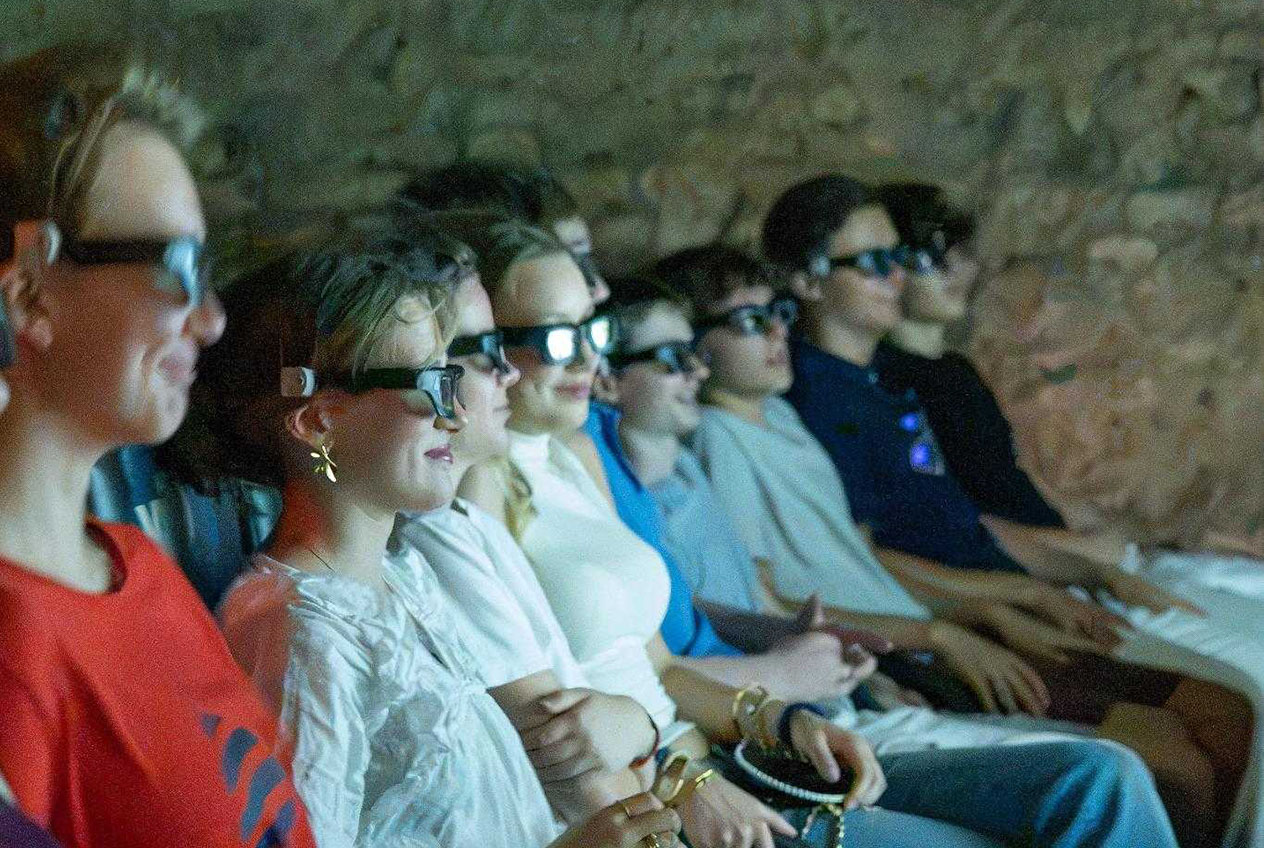 A group of people wearing 3D glasses are sitting close together in a dimly lit room, looking straight ahead and watching something just outside the frame; they appear spellbound and thoroughly entertained.