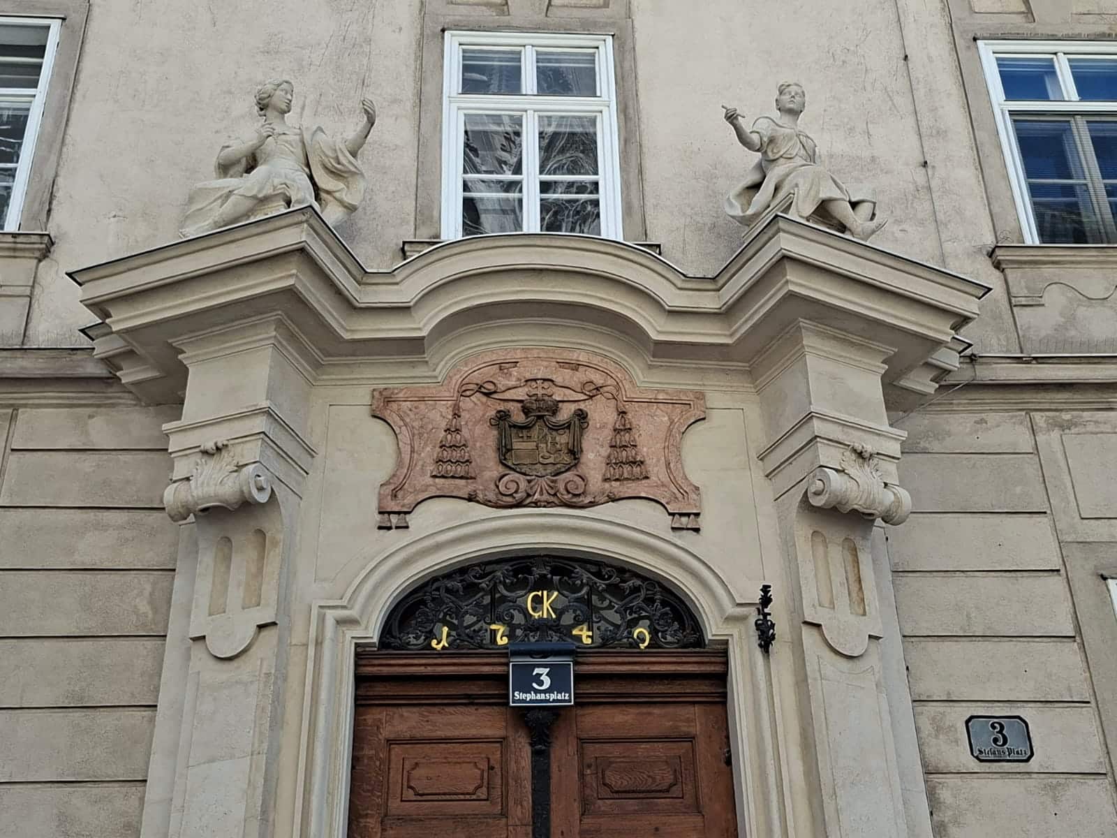 Prunkvoller Gebäudeeingang mit einem Wappen aus Stein, zwei Statuen, die ein Fenster flankieren, und dem Adressschild Stephansplatz 3 über einer hölzernen Doppeltür.