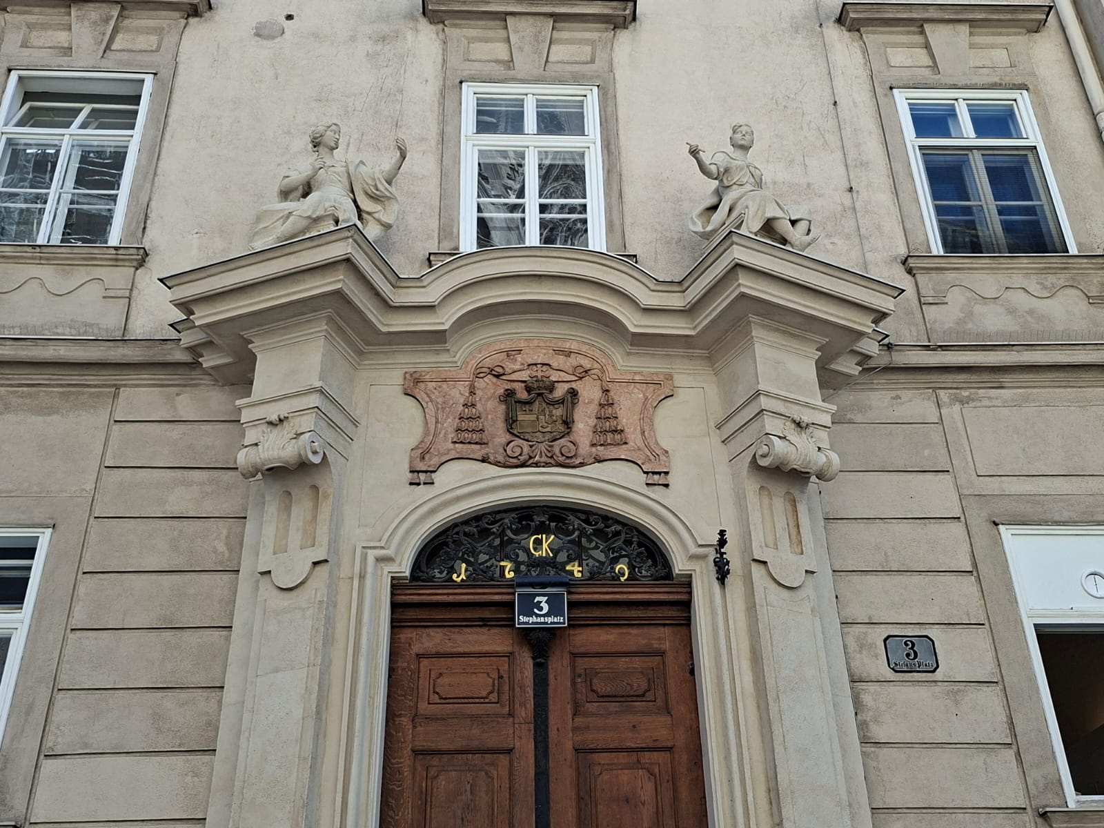 Steineingang eines Gebäudes am Stephansplatz 3 mit einer Holztür, zwei Statuen auf dem Giebel und einem verzierten Wappen.