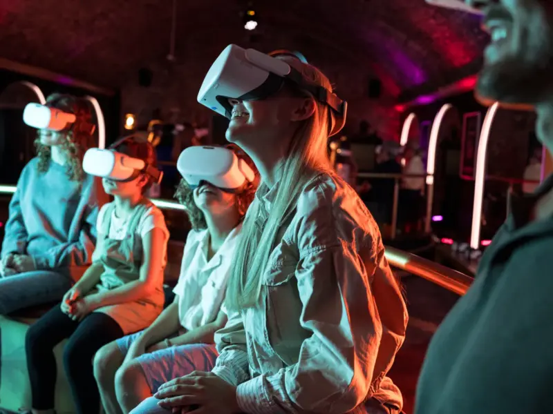 Vier Personen, die VR-Headsets tragen, sitzen in einer Reihe in einem schwach beleuchteten Raum mit violetter Stimmungsbeleuchtung.