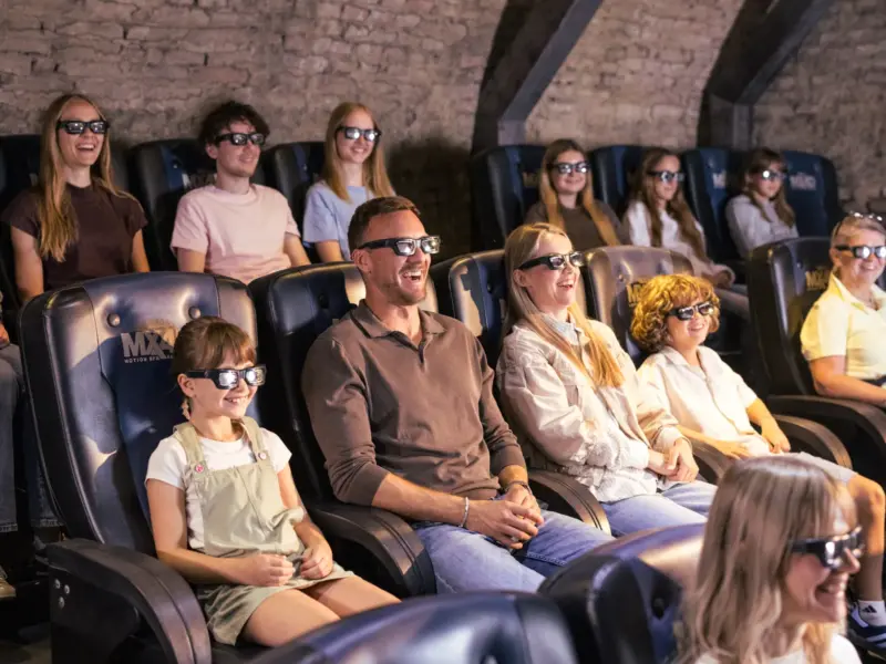 Ein Publikum mit 3D-Brillen lacht, während es in einem Kino mit Steinwänden einen Film ansieht.