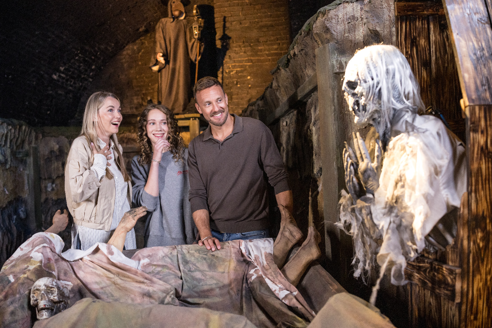 Ein Mann und zwei Frauen betrachten ein animatronisches Skelett in einer schummrig beleuchteten, kerkerartigen Attraktion.